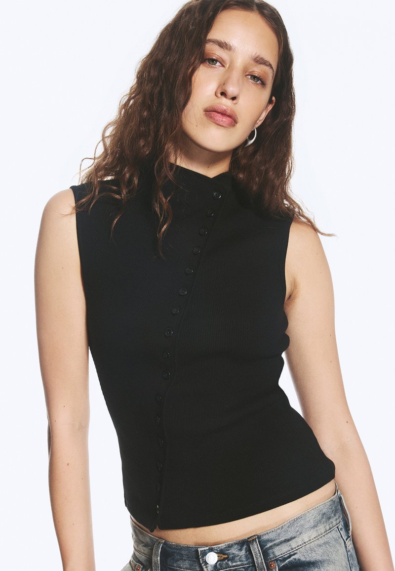 Jeune femme aux cheveux bruns ondulés portant un haut noir sans manches à boutons et un jean en denim bleu clair sur fond blanc.