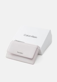 Kožený držiak na karty Calvin Klein vo svetloružovej farbe s preklopným uzáverom, striebornými kovovými prvkami a embosovaným logom, umiestnený na bielej označenej krabičke.
