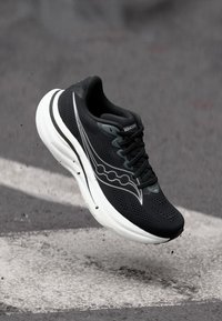 Μονόχρωμο αθλητικό παπούτσι Saucony στον αέρα επάνω από έναν ασφαλτοστρωμένο δρόμο με λευκή βαμμένη γραμμή και μικρά σωματίδια συντριμμιών από κάτω.