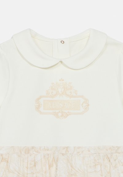 Λευκό φόρεμα με στρογγυλή γιακά, χρυσό λογότυπο "Versace" και βολάν στο τελείωμα με απαλό λουλουδένιο μοτίβο στο κάτω μέρος.