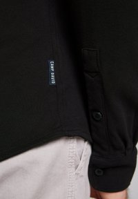 Zwarte shirt met een textuurstof, voorzien van een knoopsluiting en een 'CAMP DAVID' label aan de zijkant. Gecombineerd met lichtgekleurde broeken.