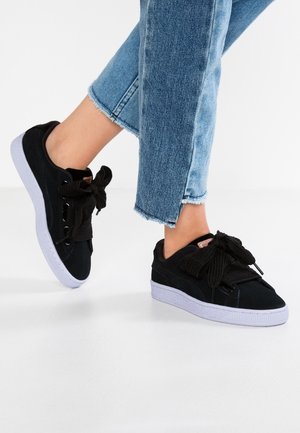 Personne portant des baskets noires avec de grands lacets en forme de nœud et un jean bleu à ourlet effiloché, debout sur un sol blanc.