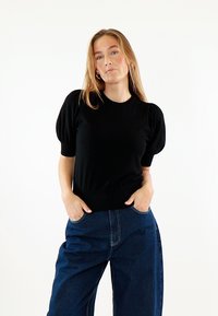 Donna con lunghi capelli biondi indossa una maglietta nera con maniche a sbuffo e jeans blu scuro a gamba larga, con le mani nelle tasche, in piedi davanti a uno sfondo bianco.