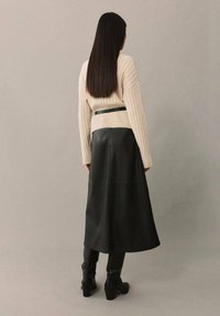 Beige geribd trui met volumineuze mouwen, gecombineerd met een zwarte leren midi-jurk en kniehoge zwarte laarzen, gezien vanuit de achterkant.