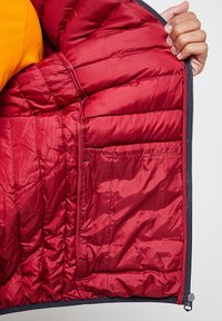 Intérieur d'une veste isolante rouge avec une texture matelassée, comportant une poche zippée et une couche de tissu orange visible en dessous.