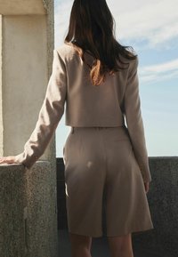 Blazer bej scurt asociat cu pantaloni scurți asortați, având cusături subtile și o textură netedă, văzut din spate.