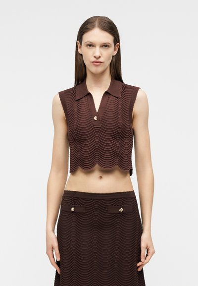 Femme portant un crop top marron texturé sans manches avec col et jupe assortie avec deux poches à boutons, debout devant un fond uni.