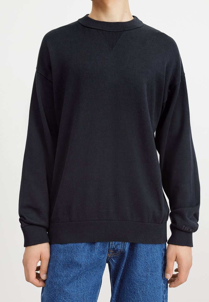 Personne portant un pull à manches longues noir uni avec col et poignets côtelés, associé à un jean en denim bleu.