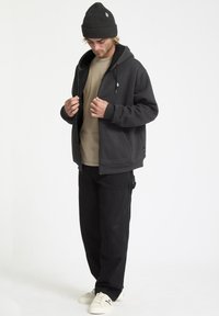 Mørkegrå zip-up hættetrøje med en blød inderside, båret over en beige t-shirt. Sammen med løse sorte bukser og hvide sneakers. Strikket sort beanie.