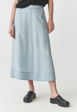 Jupe midi bleu clair portée avec un haut noir et des sabots noirs cloutés sur une personne debout devant un fond uni.