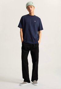 Marineblå T-shirt med korte ærmer og lille hvidt logo, kombineret med sorte bukser og ternede slip-on sko. Modellen bærer en lysegrøn hue.