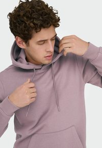 Jonge man met krullend haar die de capuchon van een mauve pullover-trui rechtzet tegen een effen lichte achtergrond.