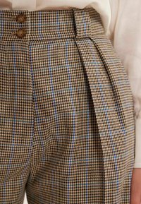 Pantalon à motif pied-de-poule marron avec des accents bleus, présentant une taille haute, des plis et deux grands boutons marron à la taille.