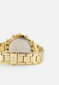 Gold-tone bracelet watch with a polished metal band, round face, chronograph functions, and a stainless steel back showing branding.