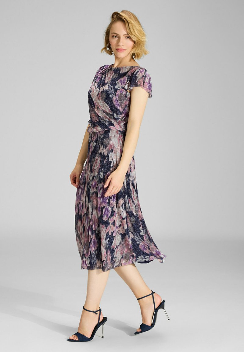 Jurk met bloemenpatroon in marineblauw, roze en crème, met een getailleerde taille en een vloeiende rok. Gecombineerd met zwarte sandalen met bandjes.