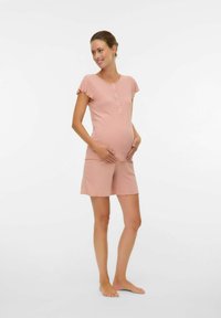 Femme enceinte en haut à manches courtes pêche et short assorti, debout pieds nus, les mains posées doucement sur son ventre, souriante et regardant sur le côté.