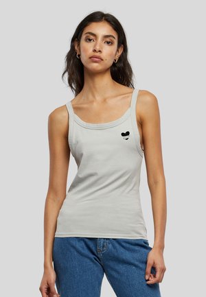 Femme portant un top sans manches gris clair avec de fines bretelles et un petit motif de cœur noir sur la poitrine, associé à un jean bleu.