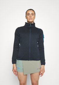 Sort fleecejakke med høj krave, lynlås foran og blå lynlåslommer. Stylet med lysegrønne shorts, der viser en tekstureret overflade.