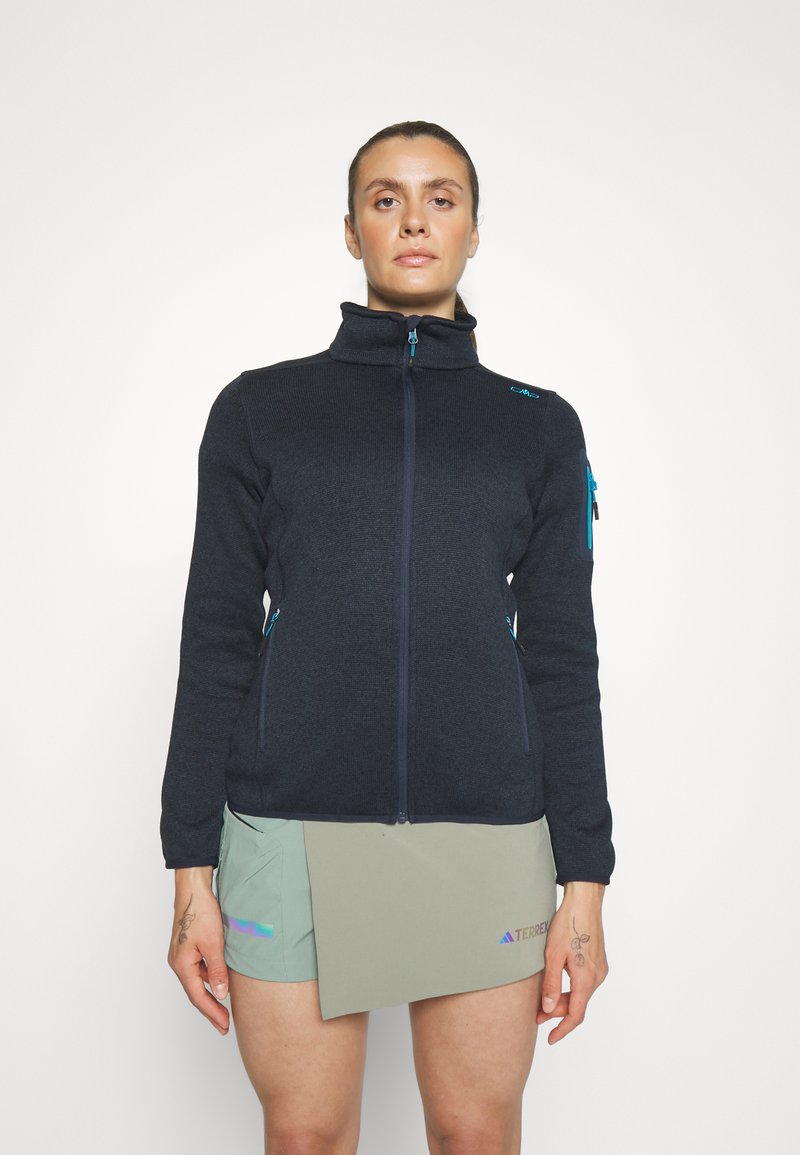 Schwarze Fleecejacke mit hohem Kragen, frontalem Reißverschluss und blauen Reißverschlusstaschen. Kombiniert mit hellgrünen Shorts, die eine strukturierte Oberfläche zeigen.