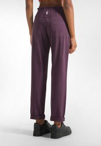 Pantaloni viola con vita elastica, orli a risvolto, due tasche posteriori e una texture liscia, abbinati a scarpe da ginnastica nere.