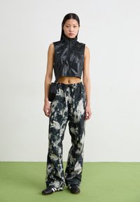 Cropped prsluk od crne umjetne kože s visokim ovratnikom, uparen s širokim hlačama u crno-bijelom tie-dye uzorku. Model drži malu crnu torbicu.