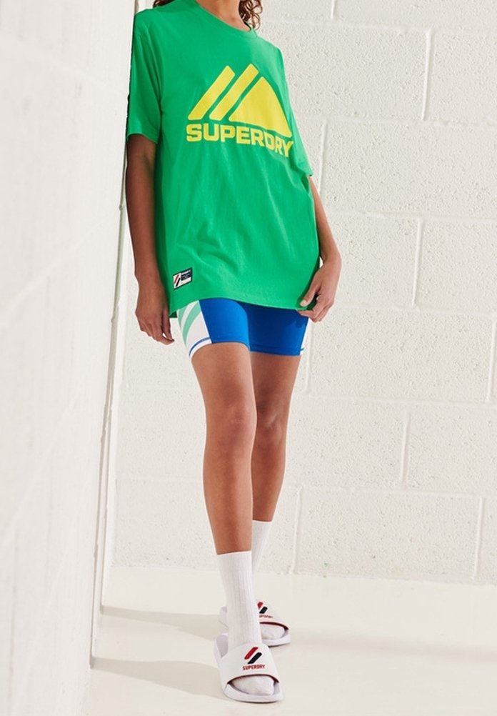 Camiseta verde con el logo amarillo de "SUPERDRY", pantalones cortos azules con acentos blancos, calcetines blancos y chanclas blancas con marca roja.