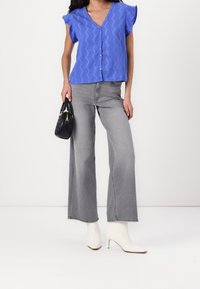 Modrá blůza s krátkým rukávem a zigzagovým vzorem, spárovaná s šedými kalhotami s vysokým pasem a širokými nohavicemi a bílými kotníkovými botami, držící černou kabelku.