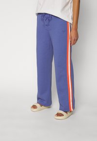 Paarse sweatpants met oranje en witte zijstrepen, een tailleband met trekkoord, en logo-detail. Gemaakt met lichtgekleurde, eenvoudige sandalen.