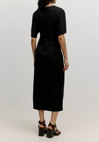 Schwarzes Midikleid mit kurzen Ärmeln, glatter Textur und seitlichem Schlitz. Kombiniert mit braunen Riemchenabsatzschuhen und Blockabsätzen.