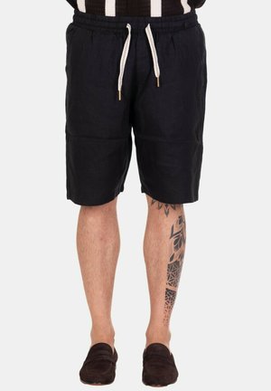 Uomo che indossa pantaloncini neri con coulisse e mocassini scuri, mostrando un tatuaggio geometrico e floreale sul polpaccio destro.