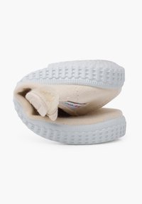 Scarpe slip-on in tela di colore crema con una superficie morbida e testurizzata, dotate di una suola in gomma blu flessibile e un'etichetta interna visibile sul tallone.
