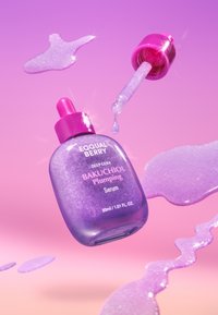 Fioletowa buteleczka serum oznaczona jako Bakuchiol Plumping z pipetą, z której wydobywa się ciecz, otoczona rozlanym, lśniącym serum na różowym tle.