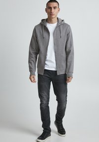 Siva zip-up dukserica s kapuljačom, nosi se preko bijele majice. Uparena s tamnim trapericama i crnim tenisicama. Glatka tkanina, ležeran dizajn.