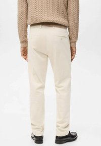 Mężczyzna ubrany w kremowe spodnie ze sztruksu, beżowy sweter z szorstkim splotem i czarne skórzane eleganckie buty, stojący na tle jednolitej białej ściany.