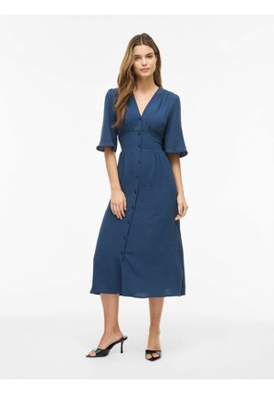 Žena v námornícky modrom midi šatách s gombíkmi vpredu a rukávmi po lakeť, v kombinácii s čiernymi otvorenými lodičkami na podpätku.