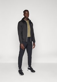 Lichte zwarte jas met rits, getailleerd olijfgroen T-shirt en zwarte tapered joggers, gecombineerd met zwarte sneakers, staande op een neutrale achtergrond.