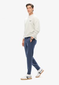 Lys grå sweatshirt med ribbede manchetter, blå sweatpants med lommer, og hvide sneakers med sorte accenter. Ensfarvet baggrund.