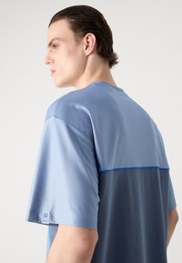 Blue short-sleeve shirt featuring a textured upper section with a smooth lower part, accented with a blue line and a small logo on the sleeve.