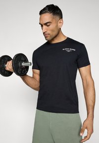 Zwarte T-shirt met ronde hals en wit logo op de linkerkant van de borst, gecombineerd met lichtgroene shorts. Het onderwerp houdt een zwarte dumbbell in de rechterhand.