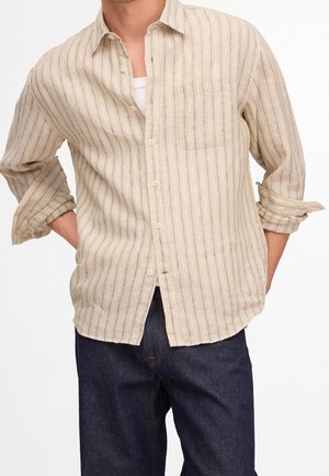 Homme portant une chemise beige à rayures boutonnée avec les manches retroussées et un jean bleu foncé, mains dans le dos.