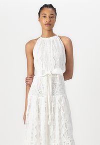 Robe blanche en dentelle avec un col halter, motif texturé, design en étages et ceinture en tissu à la taille. Liens ajustables et silhouette fluide.