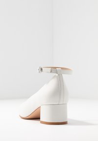 Vit konstlädersko med låg fyrkantig klack, ankelsnörning och slät yta. Har sömmar längs kanterna.