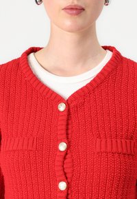 Cardigan en tricot rouge avec un col rond, des boutons dorés et une finition texturée avec des accents de côtes horizontales et verticales.