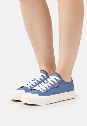 Person iført blå lave lærreds sneakers med hvide snørebånd og tykke hvide gummisåler, stående på en hvid overflade.
