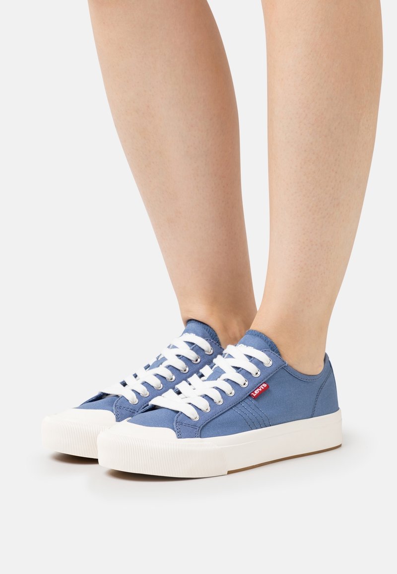 Personne portant des baskets basses en toile bleue avec des lacets blancs et des semelles épaisses en caoutchouc blanc, debout sur une surface blanche.
