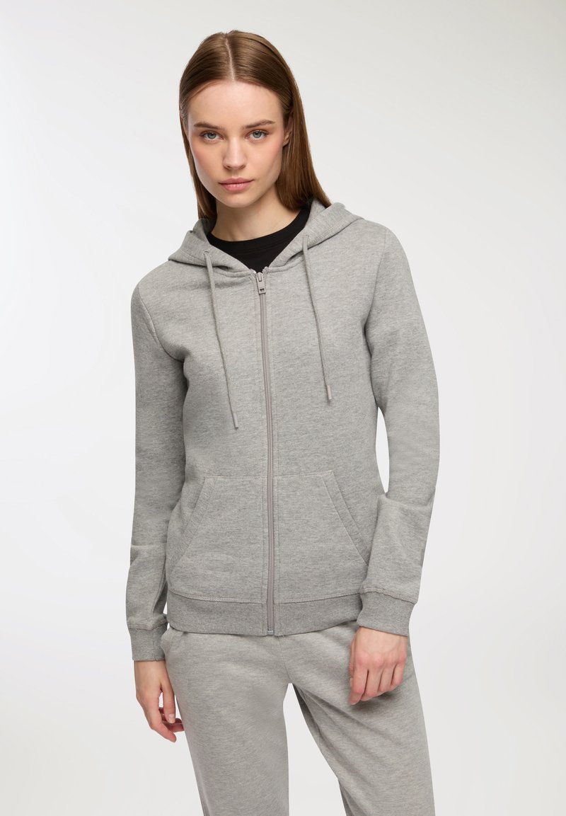 Felpa grigia con zip in tessuto morbido e testurizzato. Presenta un cappuccio, un cordoncino e tasche laterali. Indossata con pantaloni grigi abbinati.