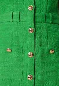 Tissu vert texturé avec de gros boutons dorés et des passants de ceinture verts assortis sur le devant d'un vêtement avec des poches.