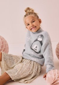Maglione fluffy azzurro chiaro con motivi a fiocco di neve, abbinato a una gonna di paillettes dorate scintillanti. Due oggetti decorativi di carta rosa nelle vicinanze.