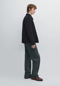 Sort jakke med en let løs pasform, båret over en hvid skjorte, parret med grå jeans og brune sko. Glat stof, minimale detaljer.