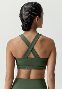 Zelená športová podprsenka s dizajnom do kríža, vyrobená z elastického materiálu. Obsahuje horizontálne švy a decentný detail loga na páske.